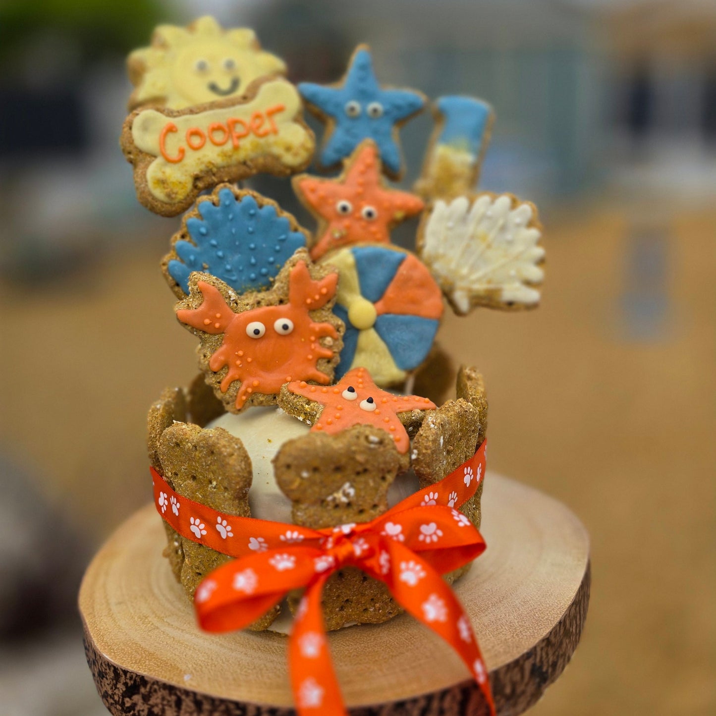decorative seaside themed cookies shaped like crabs,beach ball and shells all topped onto a bone shaped dog cake with an orange ribbon an a blurred background