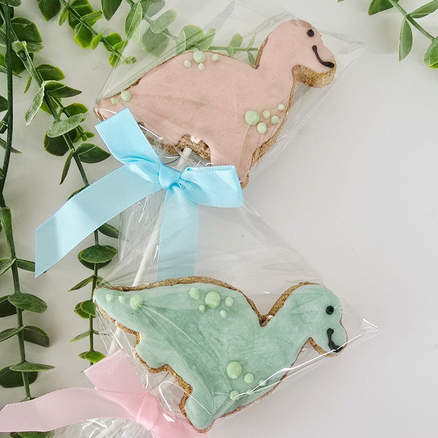 Two dinosaur shaped cookies on sticks with decorative ribbons against a white background with green leaves.
