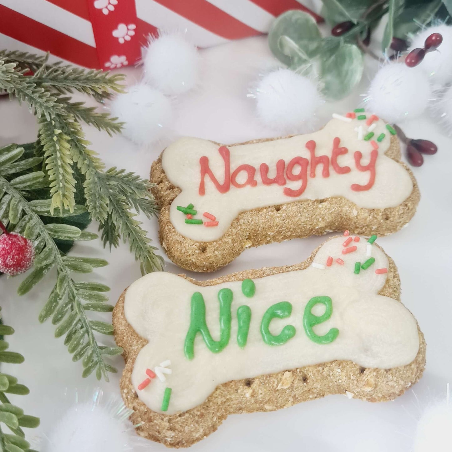 Two dog treats with the words 'Naughty' and 'Nice' written on them, placed on a decorative surface with Christmas-themed decorations around.