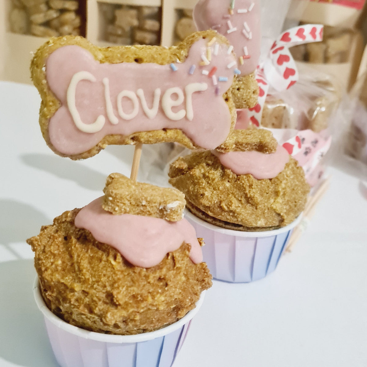 Two dog treat cupcakes with pink frosting and bone-shaped toppers reading 'Clover' with bags of dog treats blurred in the background.