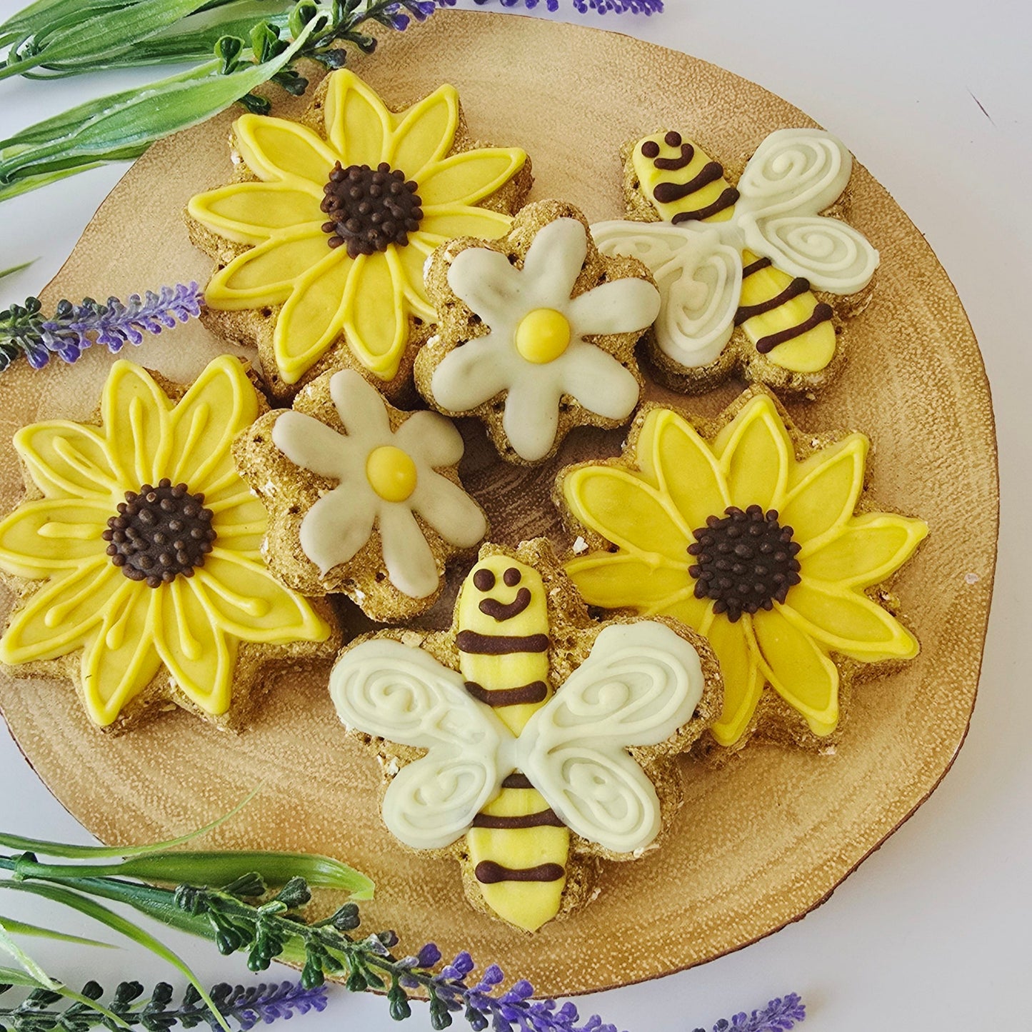 Decorative cookies shaped like bees and flowers on a wooden board with plants around.