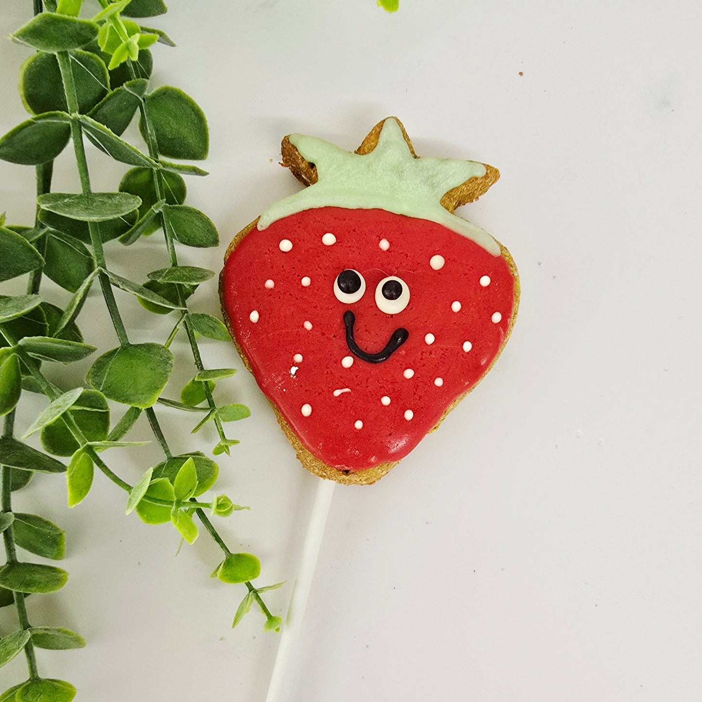 Strawberry-shaped cookie on a stick with a face, placed against a white background with green leaves.