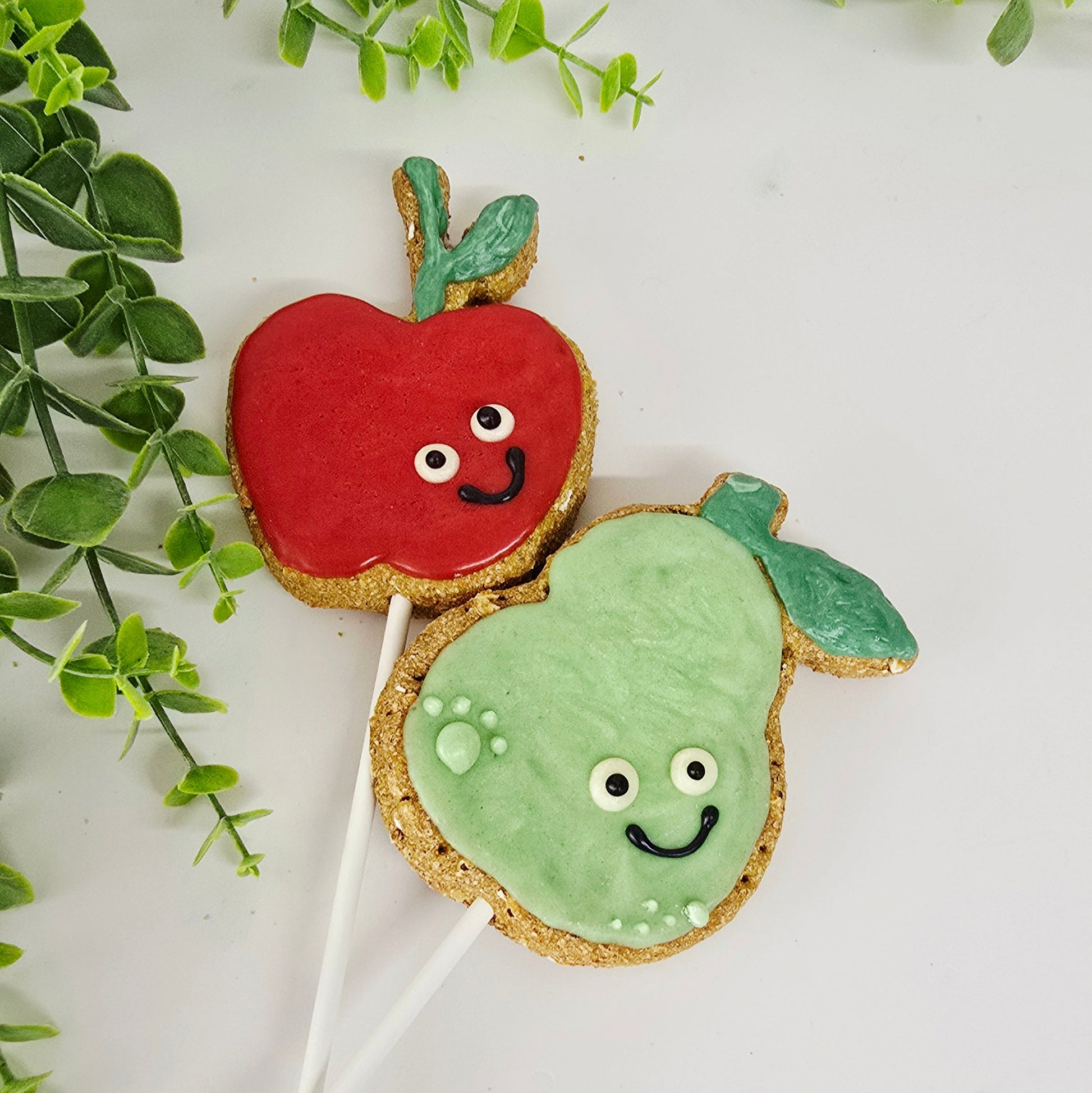 Two cookie pops 1  shaped like apple and 1 like a pear  both with faces on a white background with green leaves