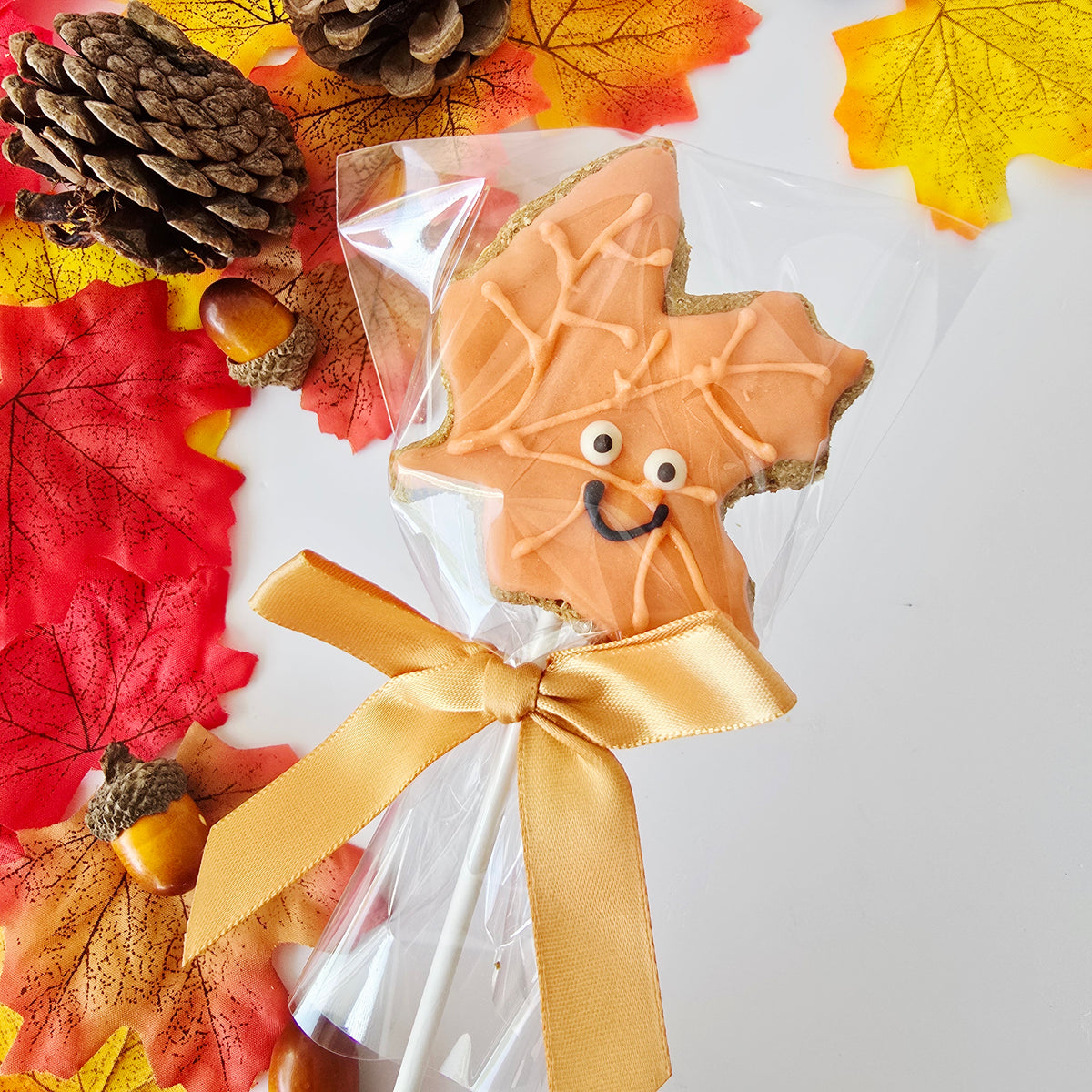 An autumn-themed cookie lollipop featuring a maple leaf-shaped cookie with orange icing, web patterns, and a smiling face, wrapped in a clear bag with a gold ribbon bow. The product is styled with fall leaves, pinecones, and acorns on a white background.