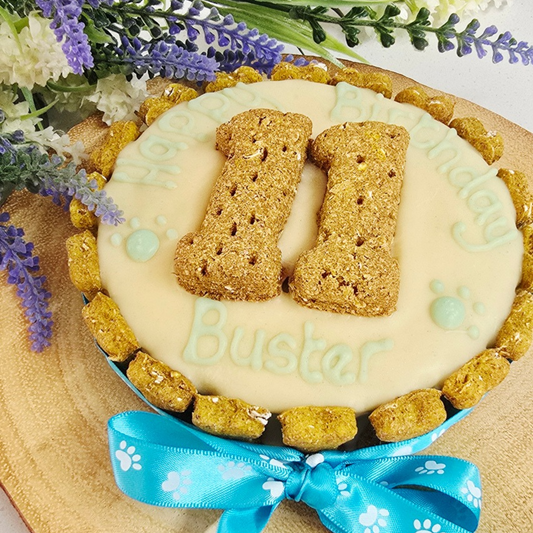A round pet birthday cake decorated with pale cream dog friendly icing, topped with number shaped biscuits, the name 'Buster' in carob icing, paw prints, and a blue ribbon bow at the bottom. The cake is photographed on a wooden surface with flowers in the background.