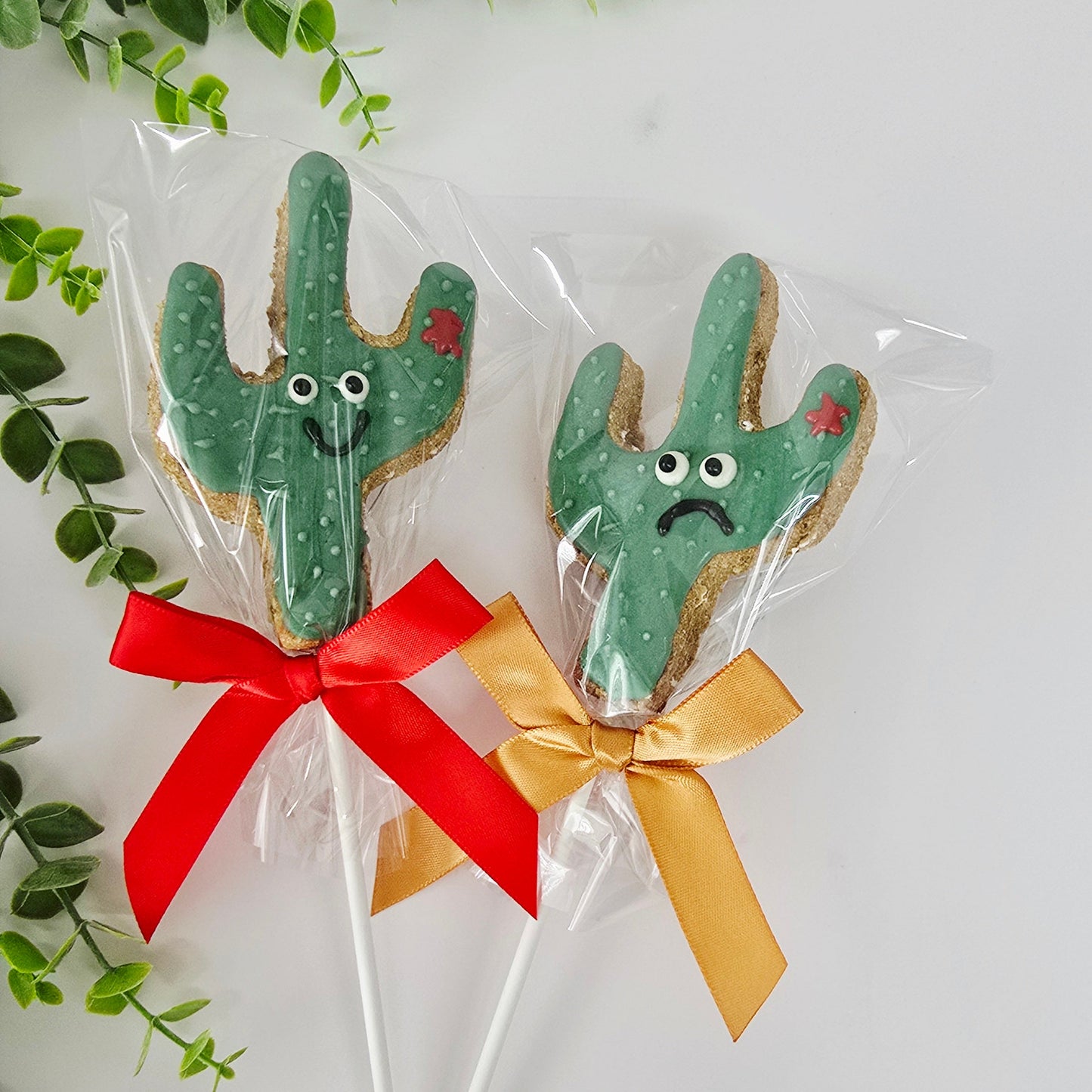 Two cactus-shaped cookies on sticks with red and gold bows, surrounded by green leaves.