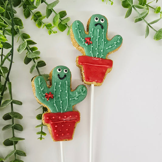 Two cactus-shaped cookies on sticks with green leaves and red flowers, surrounded by greenery on a white background.