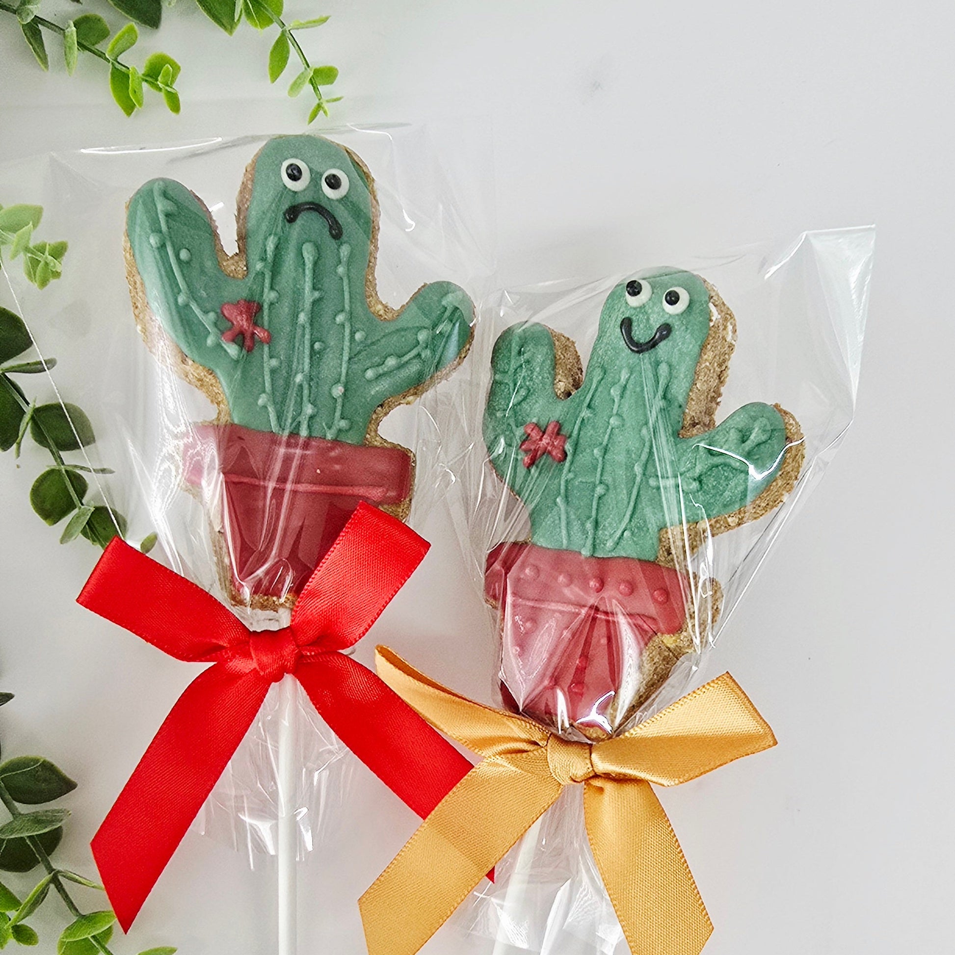 Two cactus-shaped cookies on sticks with red and gold ribbons, surrounded by green leaves.