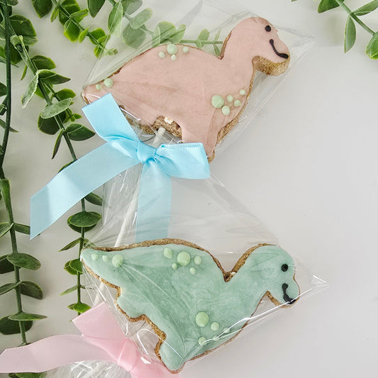 Two dinosaur shaped cookies on sticks with decorative ribbons against a white background with green leaves.