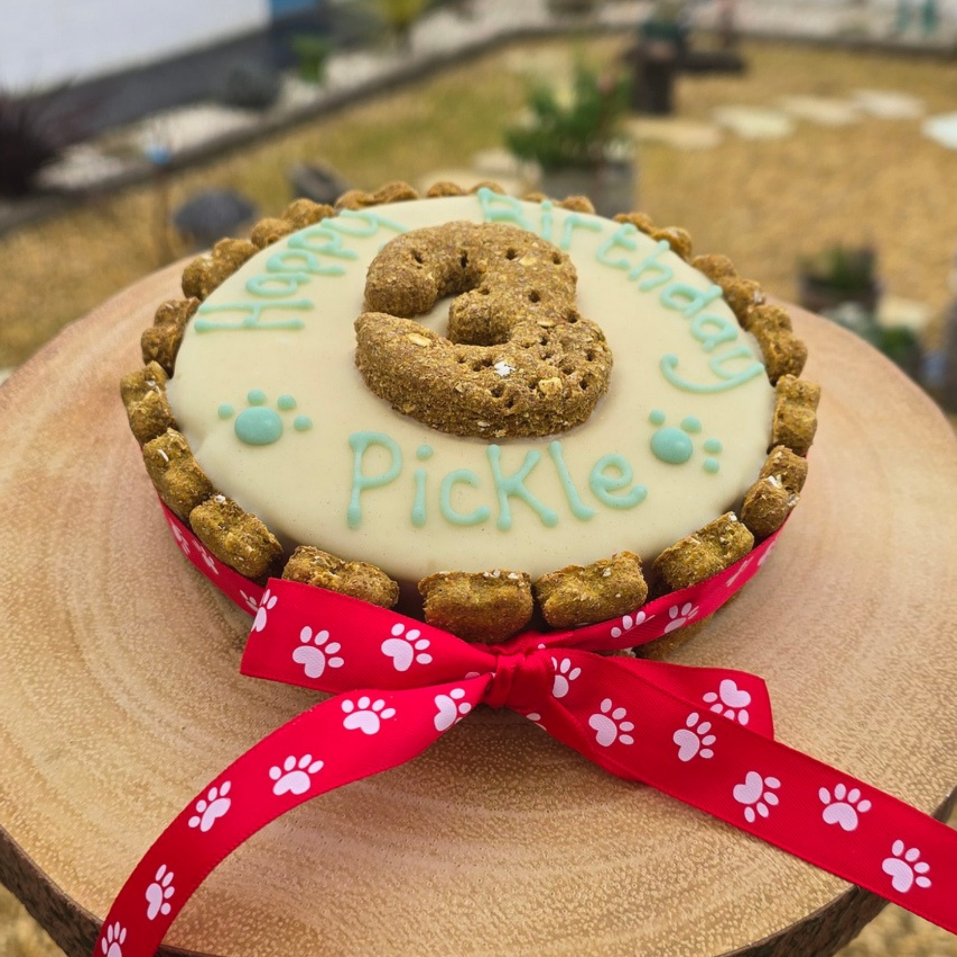 A round dog treat cake decorated with white frosting, blue icing text reading 'Happy Birthday Pickle,' a biscuit cookie number 3 on top, and a red ribbon with white paw prints tied around the base, shown on a wooden slice outdoors.Handmade decorated dog friendly birthday cake custom to your dog .