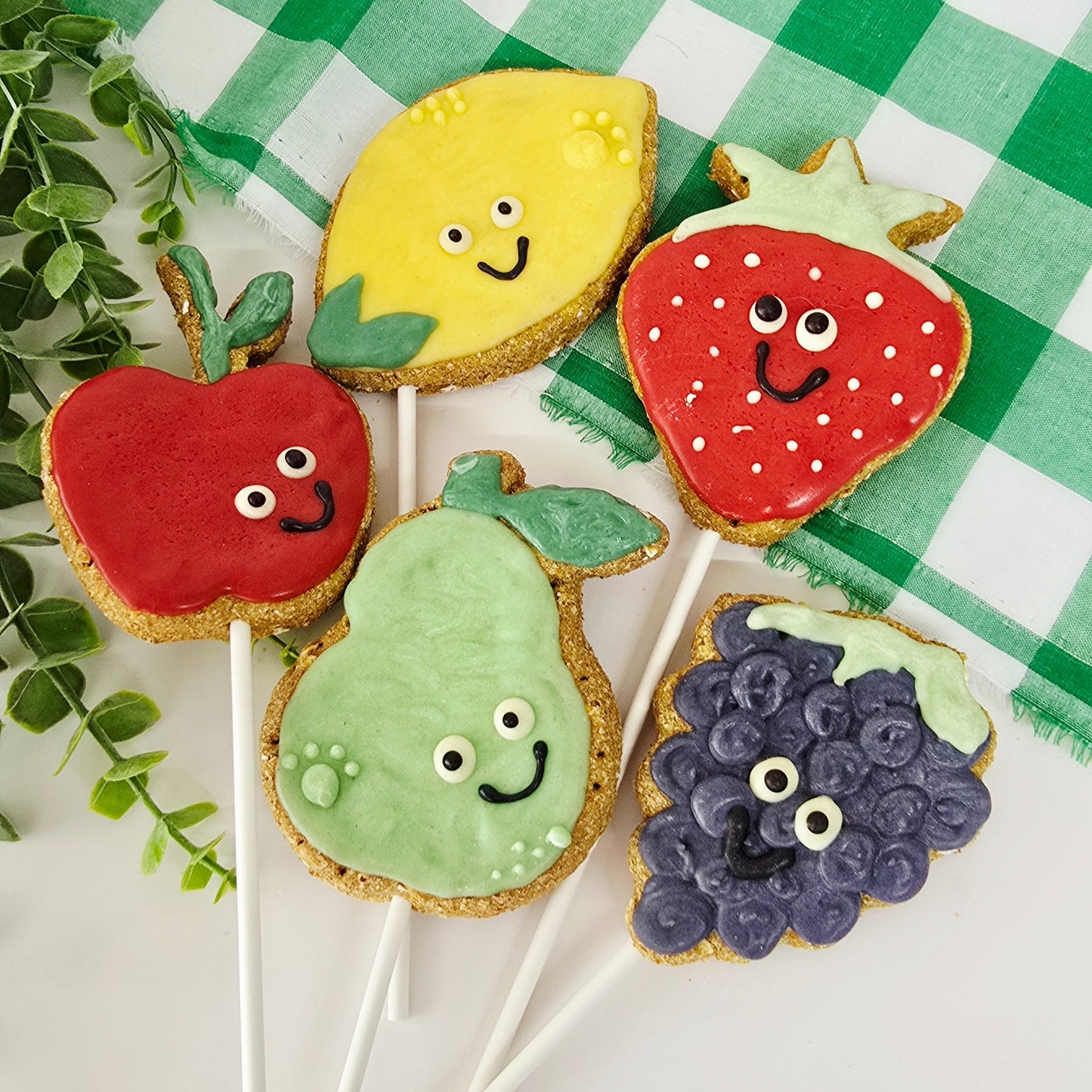 Colorful cookies shaped like fruits  on sticks with a green checkered tablecloth.