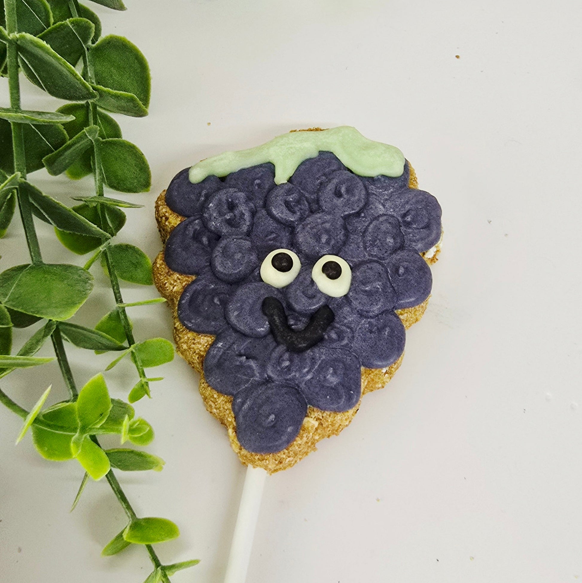 Decorative  grape lollipop shaped like  with purple accents and a smiley faceon a white background with green leaves.