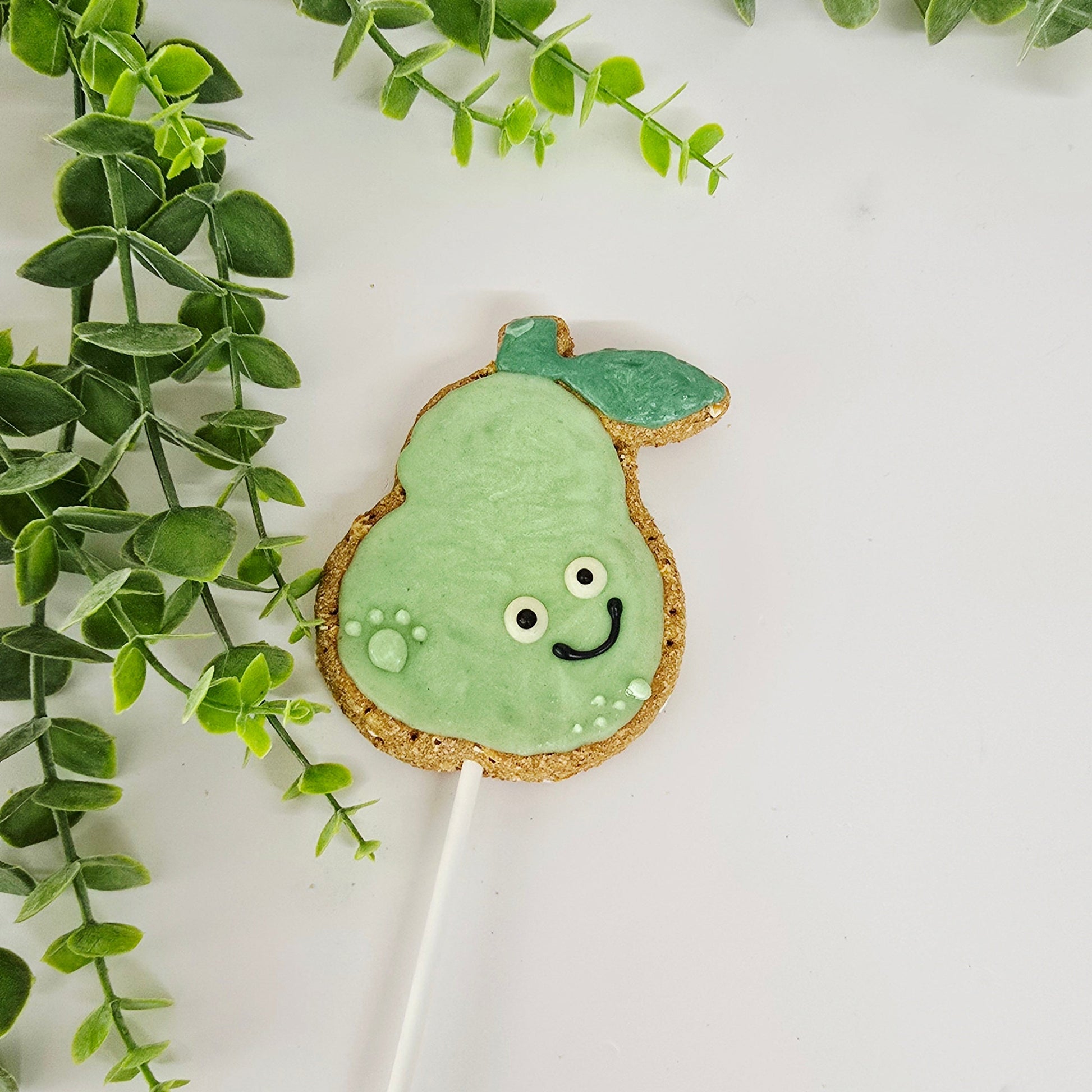 Green pear cookie on a stick with a face, surrounded by green leaves on a white background
