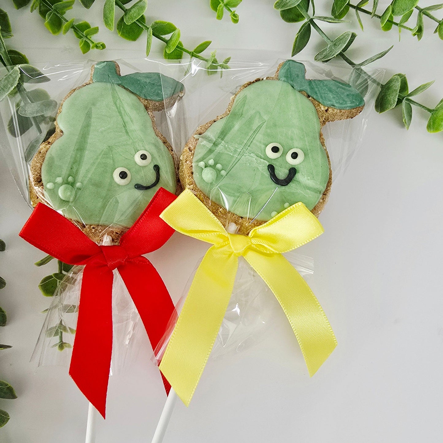 Two green cookie pops with faces on sticks, one with a red bow and the other with a yellow bow, surrounded by green leaves.