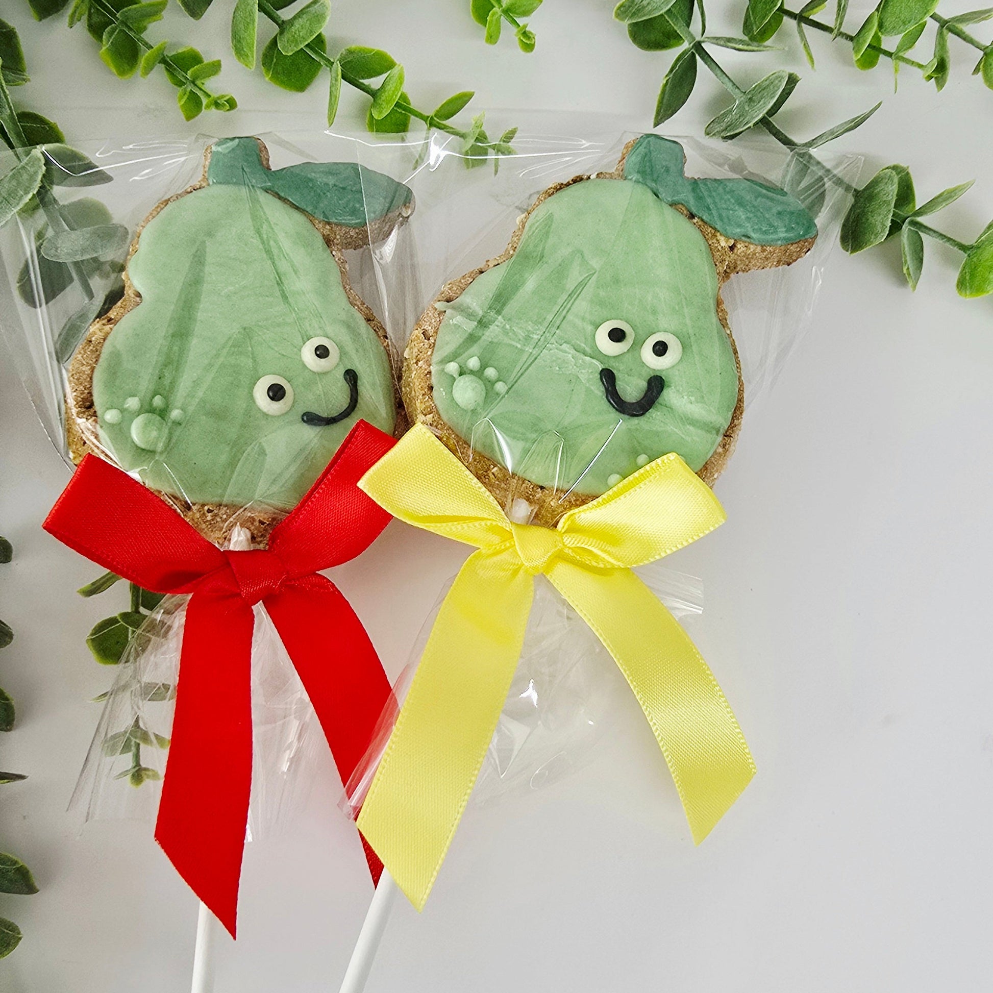 Two green cookie pops with faces on sticks, one with a red bow and the other with a yellow bow, surrounded by green leaves.