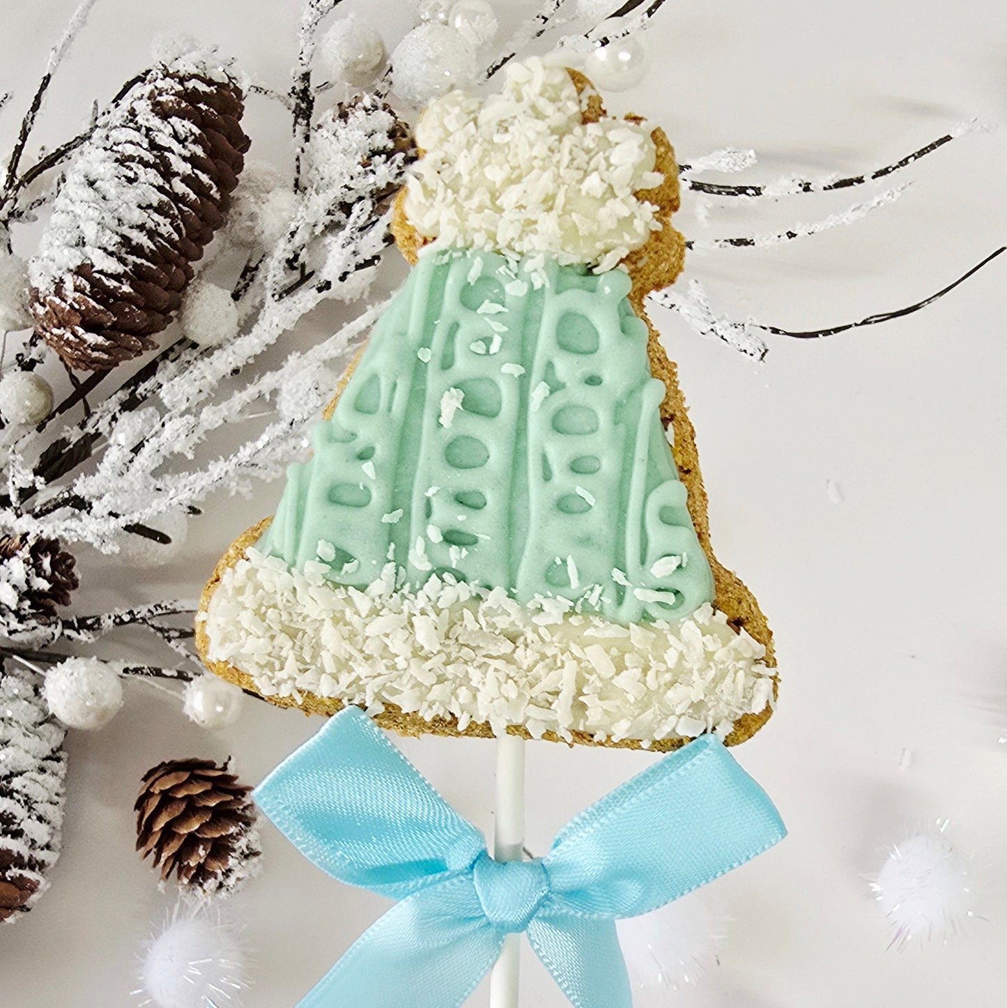 Decorative cookie on a stick with a green dress and white flowers, tied with a blue ribbon, against a snowy branch background.