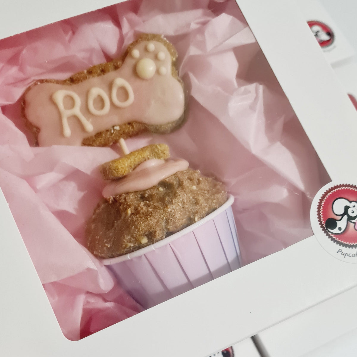 A hand holds a white gift box with a clear window, showing a decorated dog treat gift set with a cookie shaped like a paw print reading 'Roo' and a pink cupcake in a striped paper liner, lined with pink tissue paper.