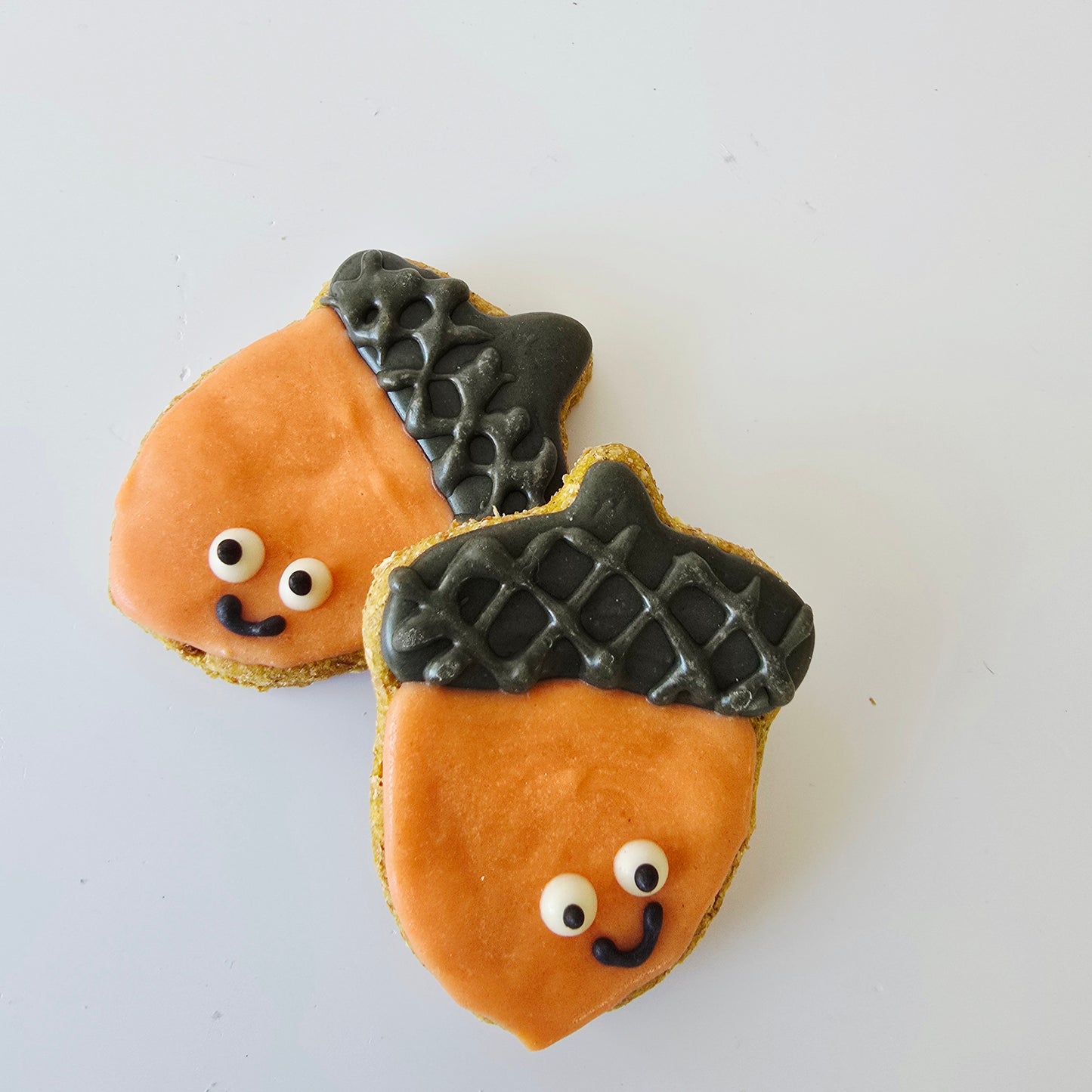 Two acorn-shaped cookies decorated with orange carob for dogs, brown textured dog friendly icing on top, and cute white candy eyes with black smiles, arranged on a plain white background.