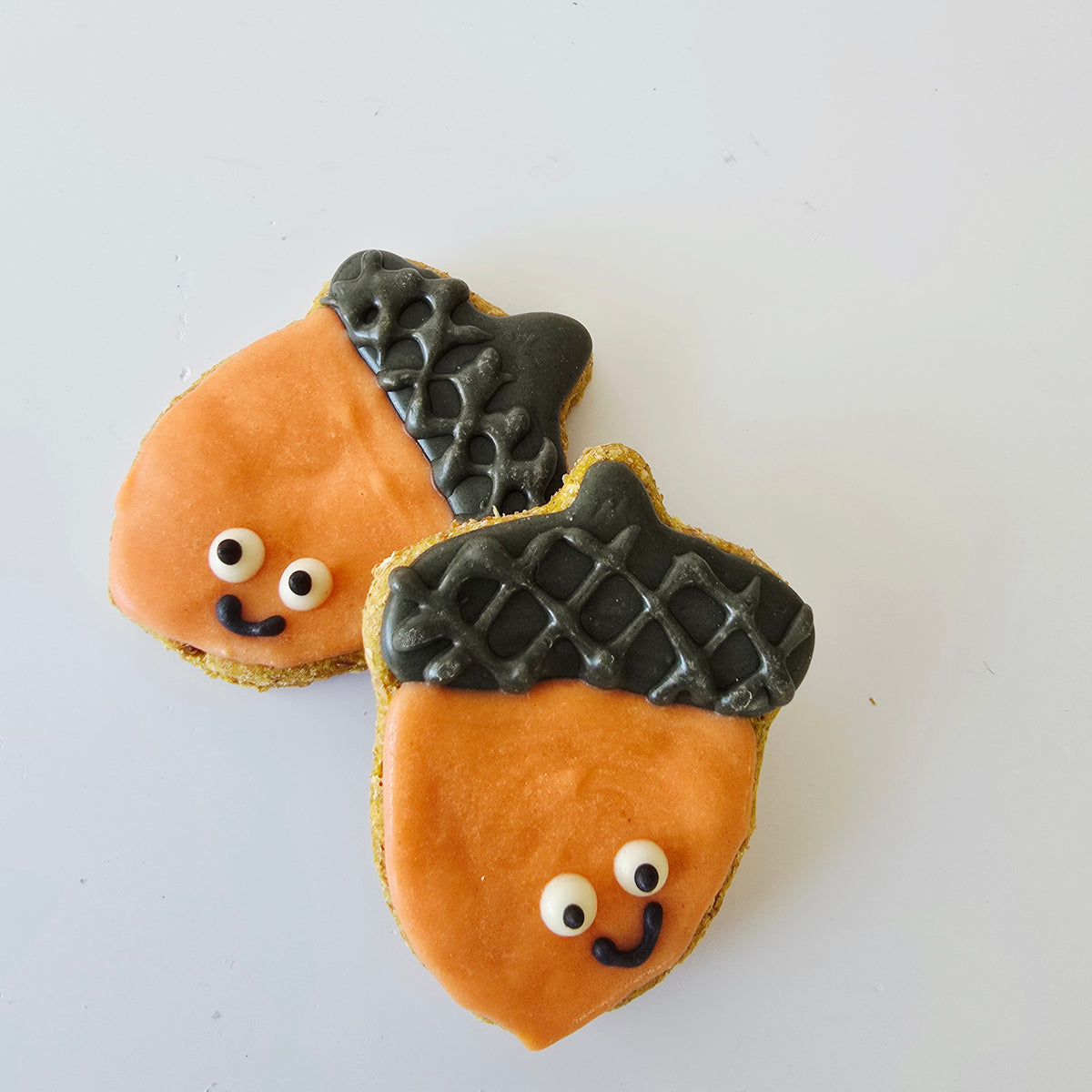 Two acorn-shaped cookies decorated with orange carob for dogs, brown textured dog friendly icing on top, and cute white candy eyes with black smiles, arranged on a plain white background.