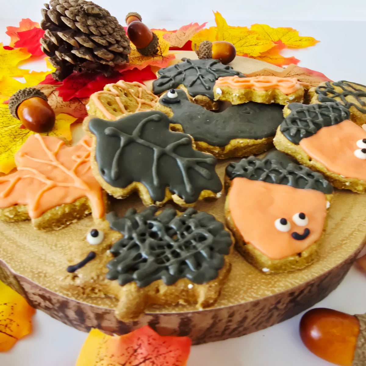 Autumn-themed dog treat cookies decorated as orange acorns, brown hedgehogs, and maple leaves, arranged on a rustic wood slice with fall leaves, acorns, and a pinecone.