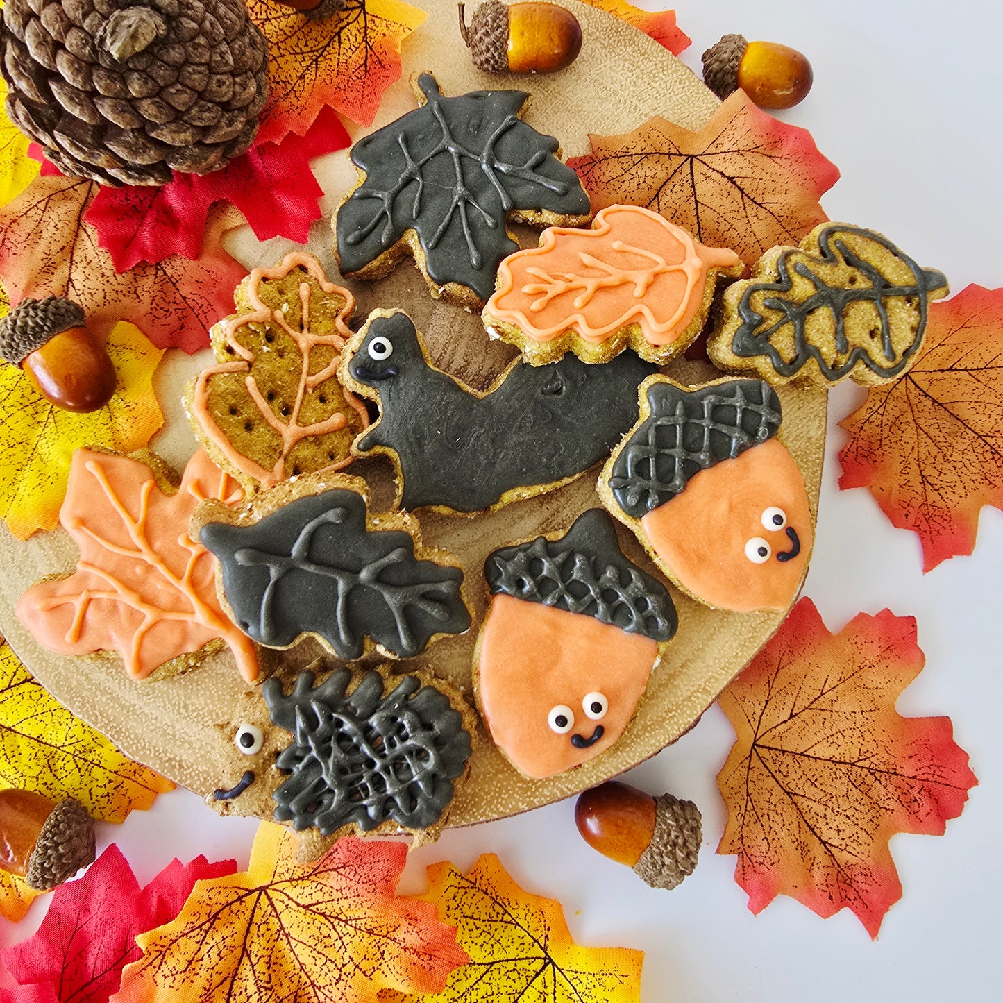 Decorated autumn-themed dog  friendly cookies shaped like leaves, acorns, a bat, and a hedgehog, arranged on a round wooden board with fall leaves, acorns, and a pinecone around them.