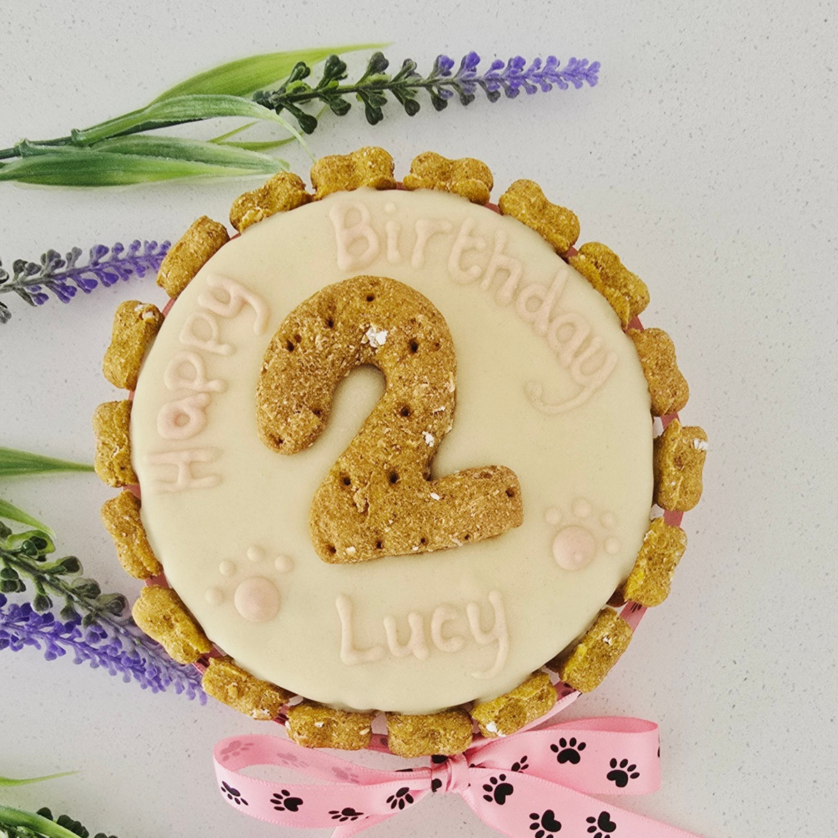 A round pet treat cake decorated with white icing, a large cookie number 2 in the center, the words 'Happy Birthday Lucy' in icing, and a pink ribbon tied in a bow at the bottom. The cake is topped with handmade golden-brown biscuit bones around the edge and is photographed on a light surface with decorative green and purple flowers in the corners.All handmade and dog friendly