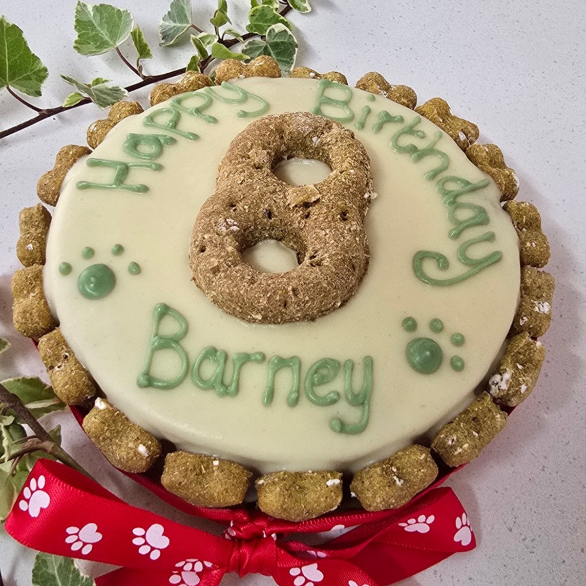 A round dog treat birthday cake decorated with dog friendly icing, green 'Happy Birthday Barney' icing, a large cookie number 8, and a red paw-print ribbon bow at the bottom. The cake is topped with dog treats around the edge and placed on a light surface with green leaves in the corner.All is handmade and personalised 