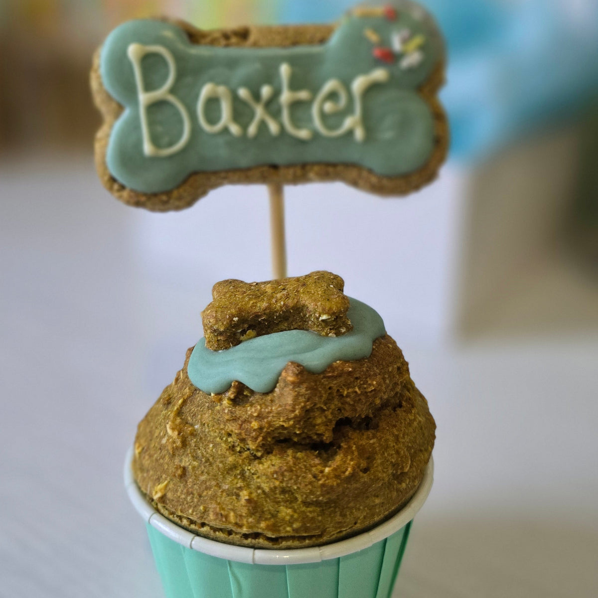 A personalised cupcake featuring a golden-brown baked dog treat on top with light blue frosting, a light blue cupcake liner, and a bone-shaped cookie topper with the name 'Baxter' in white icing.