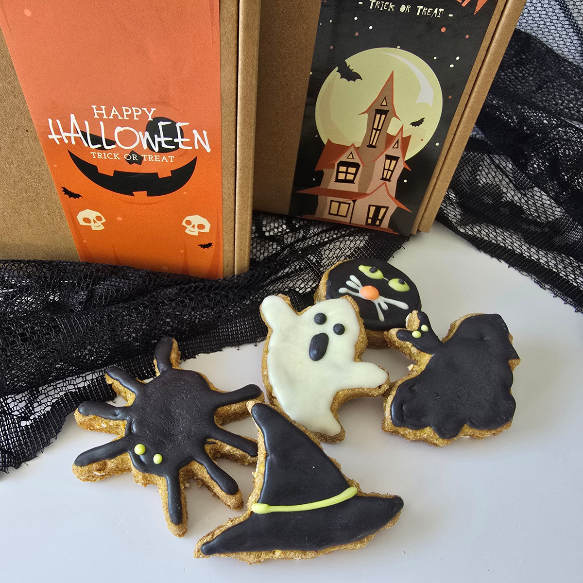 Five Halloween-themed dog friendly cookies shaped like a spider, ghost, witch hat, and cat, arranged on a white surface in front of a Halloween gift box and black lace fabric.