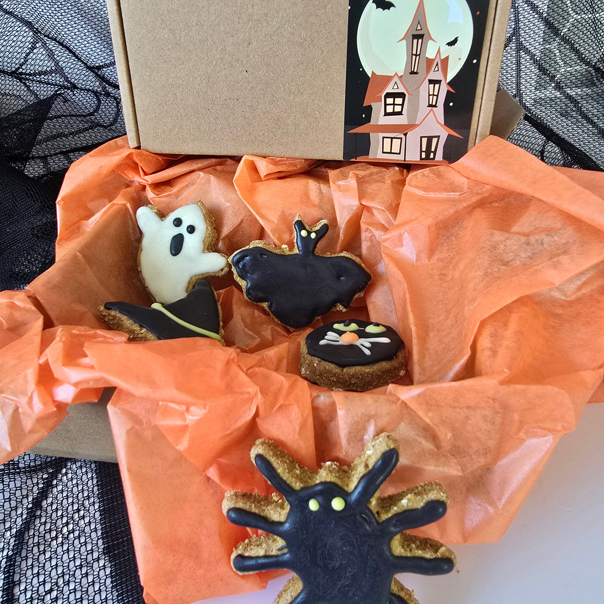 Halloween-themed dog cookie box filled with orange tissue paper, featuring four decorated cookies shaped like a ghost, bat, cat, and spider, with a brown cardboard box in the background.