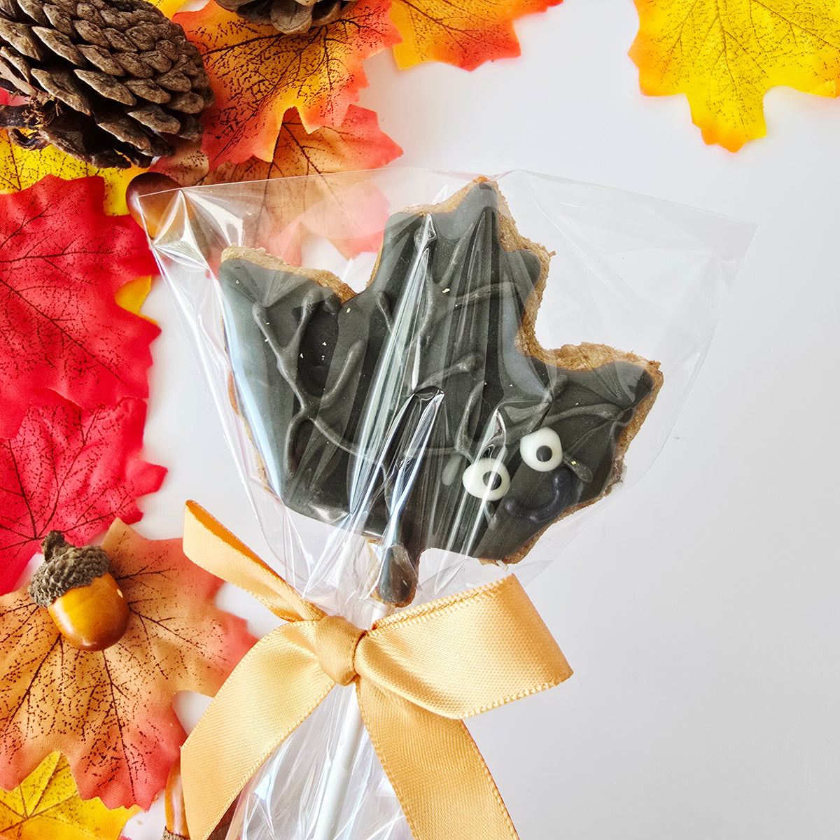 An Autumn wimter -themed leaf-shaped cookie wrapped in clear cellophane and tied with a gold ribbon,onto a paper stick  displayed on a white background with autumn leaves, a pinecone, and an acorn.