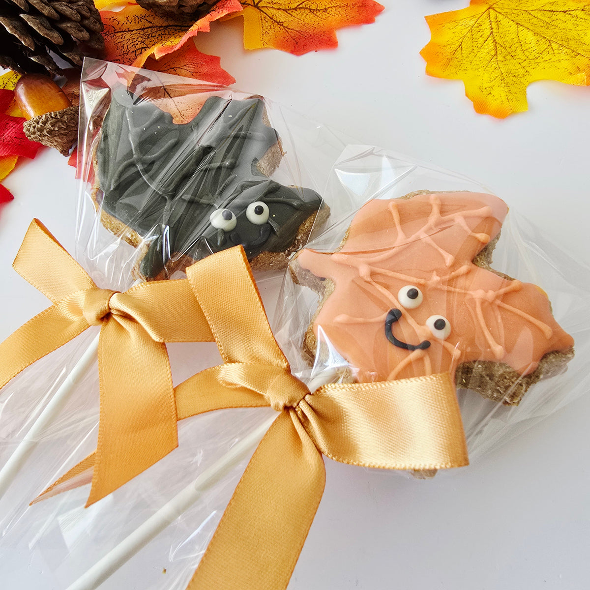 Two Halloween-themed bat-shaped cookies in clear cellophane bags with gold ribbon laces, decorated with orange and dark gray icing and candy eyes, on a white background with autumn leaves.