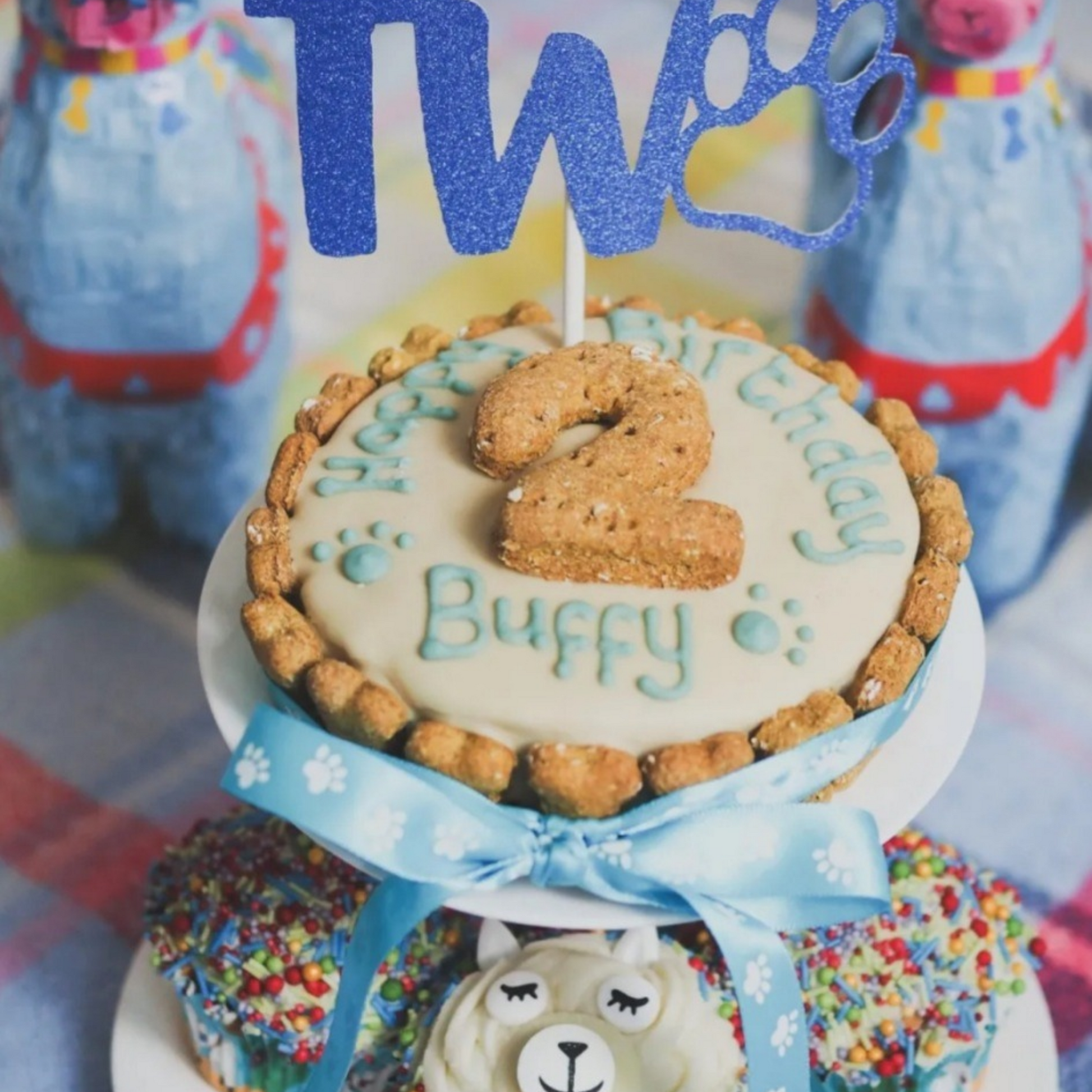 A custom pet birthday cake on a white tiered stand, decorated with beige frosting, blue piped lettering that reads 'Happy Birthday Buffy', a blue ribbon, and a blue glitter 'Two' topper. The cake is topped with a cookie-shaped number '2' candle.