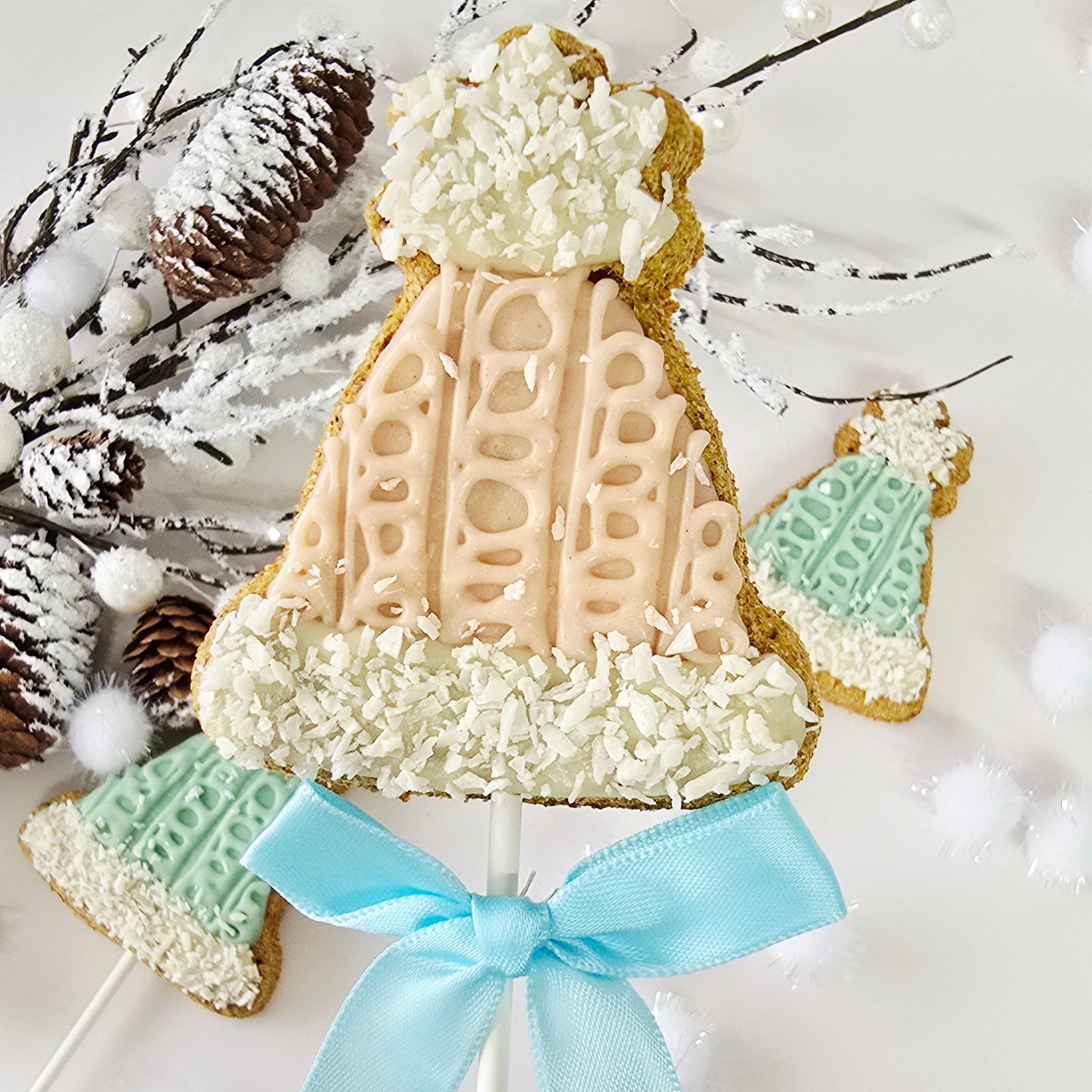 Decorative cookie on a stick shaped like a sweater with a blue ribbon, surrounded by pine cones and branches.
