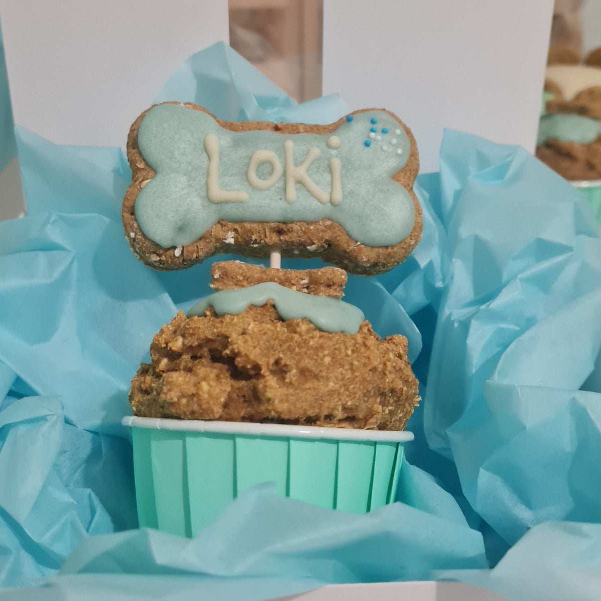 A custom doggie  cupcake in a mint green liner, decorated with a light blue icing topper shaped like a bone with the name 'Loki' in icing, nestled in light blue tissue paper inside a white box.Handmade and personalised for the dog
