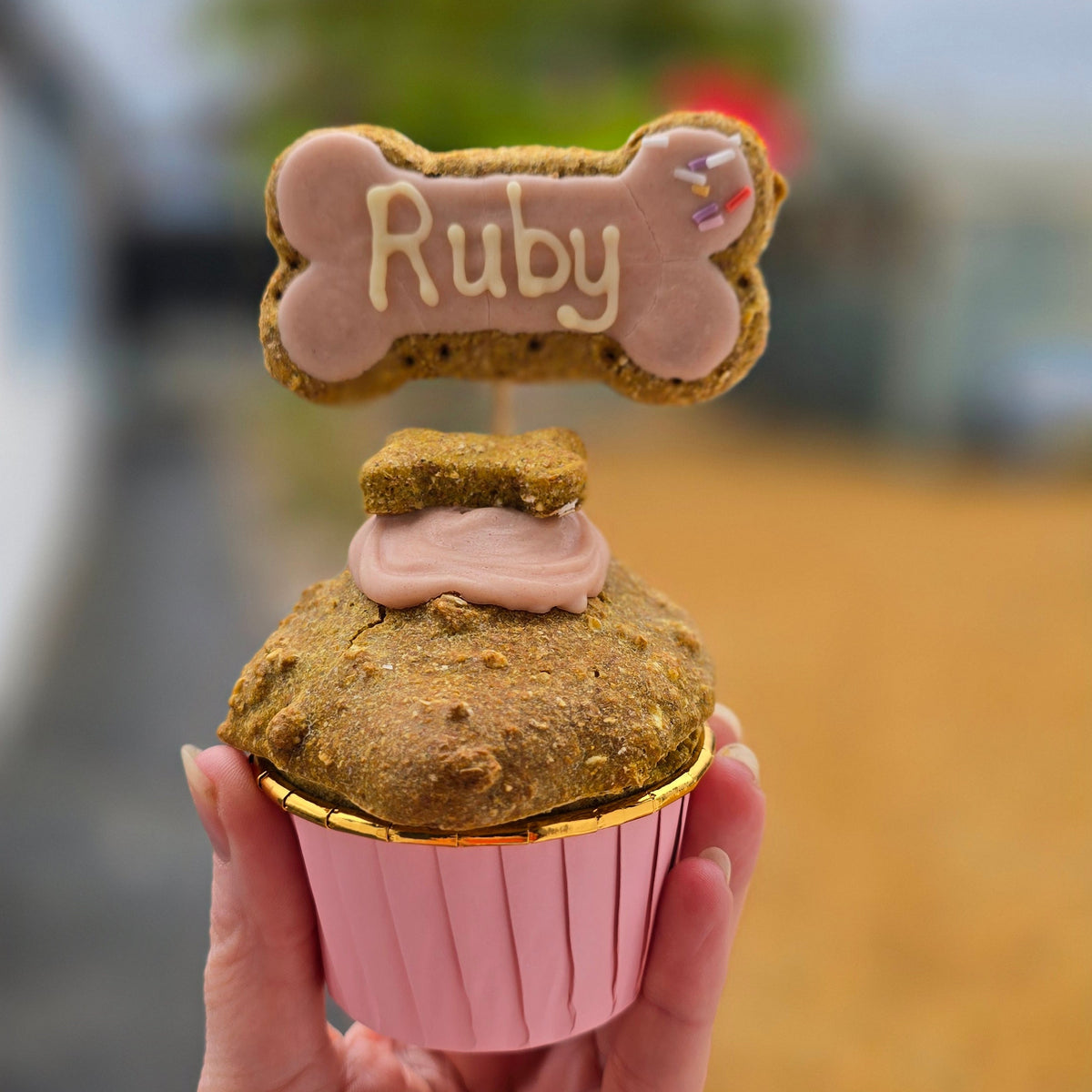 A hand holds a decorated dog treat cupcake outdoors. The cupcake has pink frosting, a pink paper liner, and a bone-shaped topper that says 'Ruby' in icing.