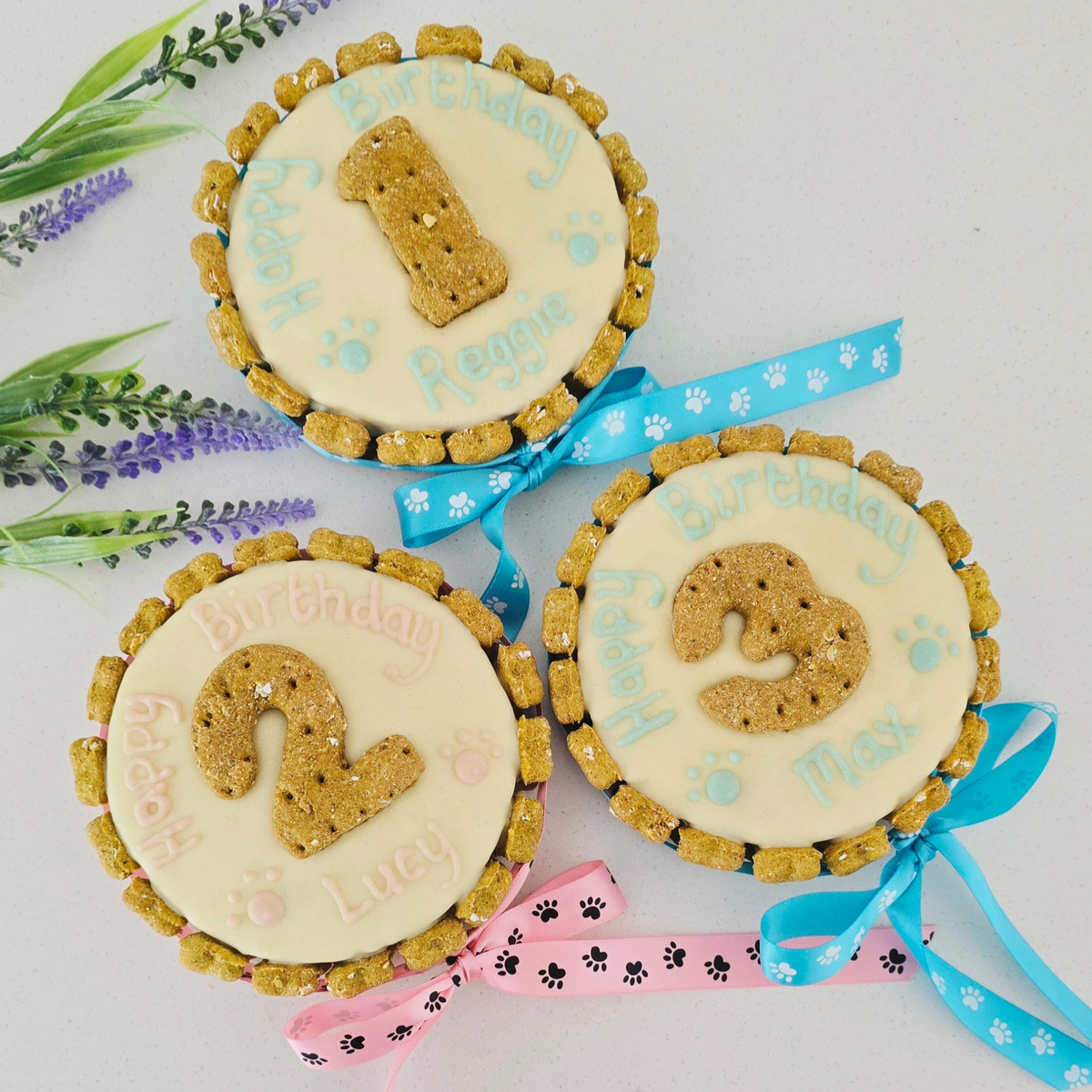 Three round dog cakes decorated with custom birthday frosting and numbered 1, 2, and 3, tied with pink, blue, and light blue ribbons, arranged on a white background with decorative flowers.Dog friendly icing and no peanut butter 