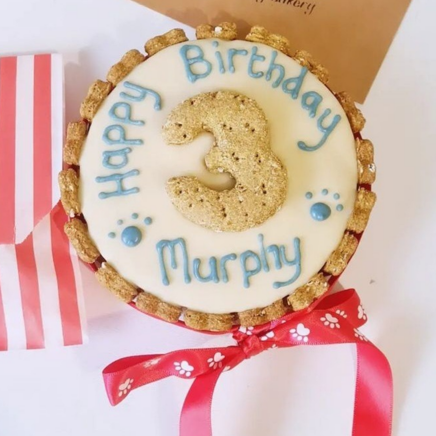 A round decorated dog friendly  birthday  cake  with cream carob icing, blue text reading 'Happy Birthday Murphy' and a large number 3, shown with a red ribbon and a striped gift box on a white background.