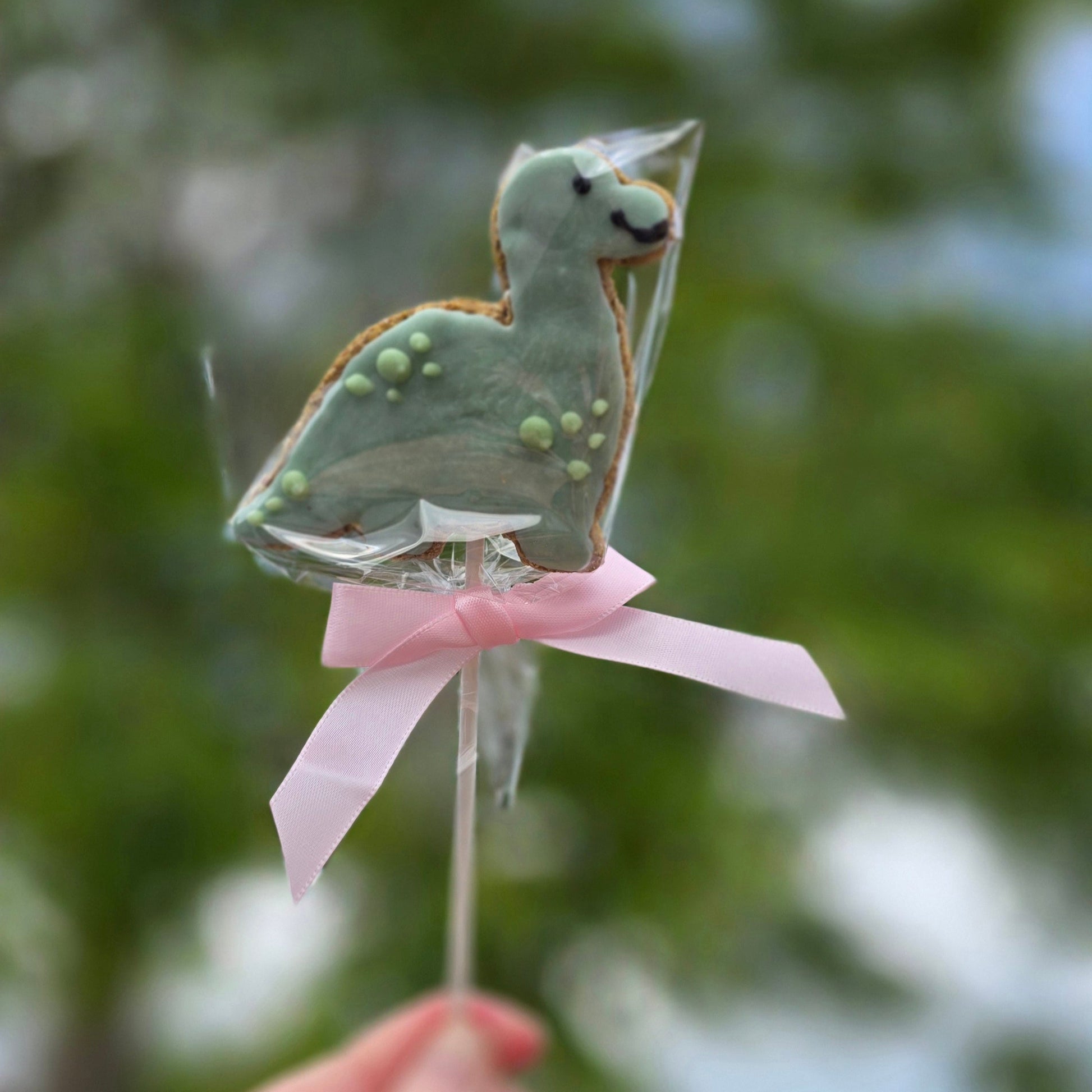 Blue dinosaur-shaped lollipop on a stick with a pink ribbon held against a blurred green background.
