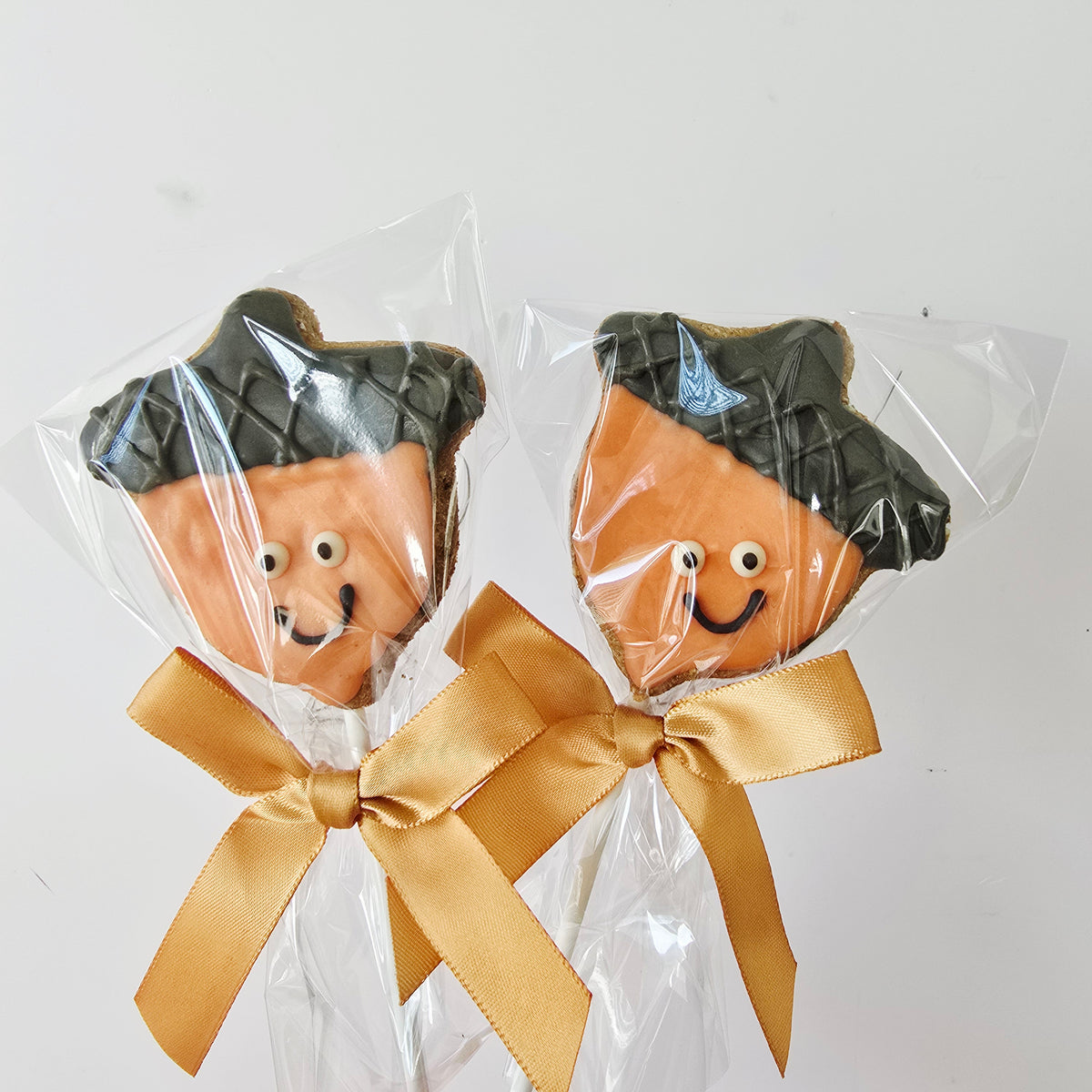 Two acorn-shaped lollipops decorated with orange-brown icing and black acorn caps, wrapped in clear cellophane with gold ribbon bows, on a white background.