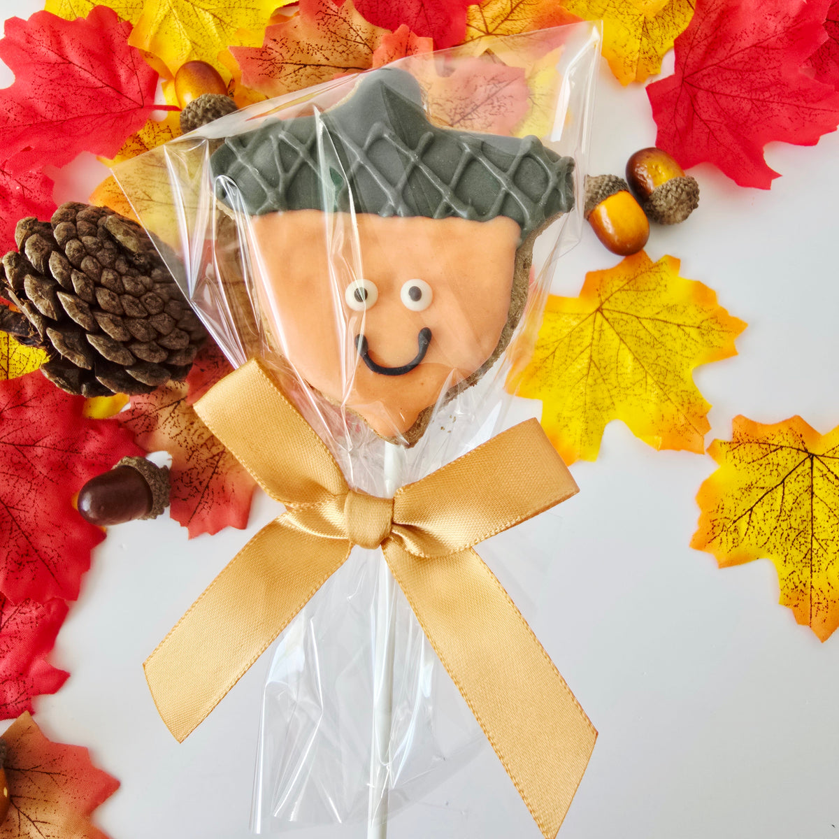 Acorn-shaped decorated cookie on a stick, wrapped in clear cellophane with a gold ribbon bow, surrounded by autumn maple leaves, acorns, and a pine cone on a white background.