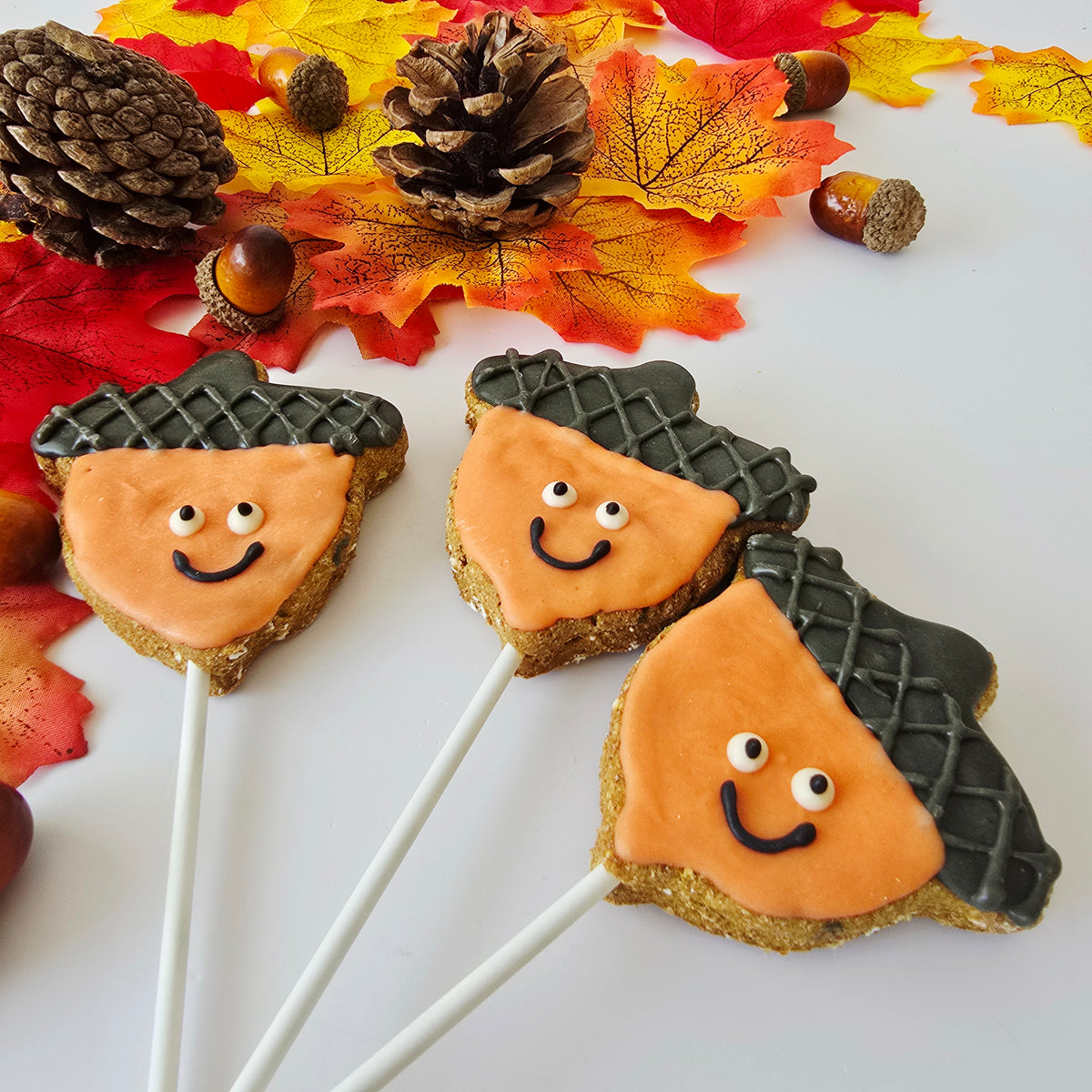 Three acorn-shaped sugar cookies with orange icing faces and dark gray icing acorns on white sticks, styled with autumn leaves, pinecones, and acorns on a white surface.