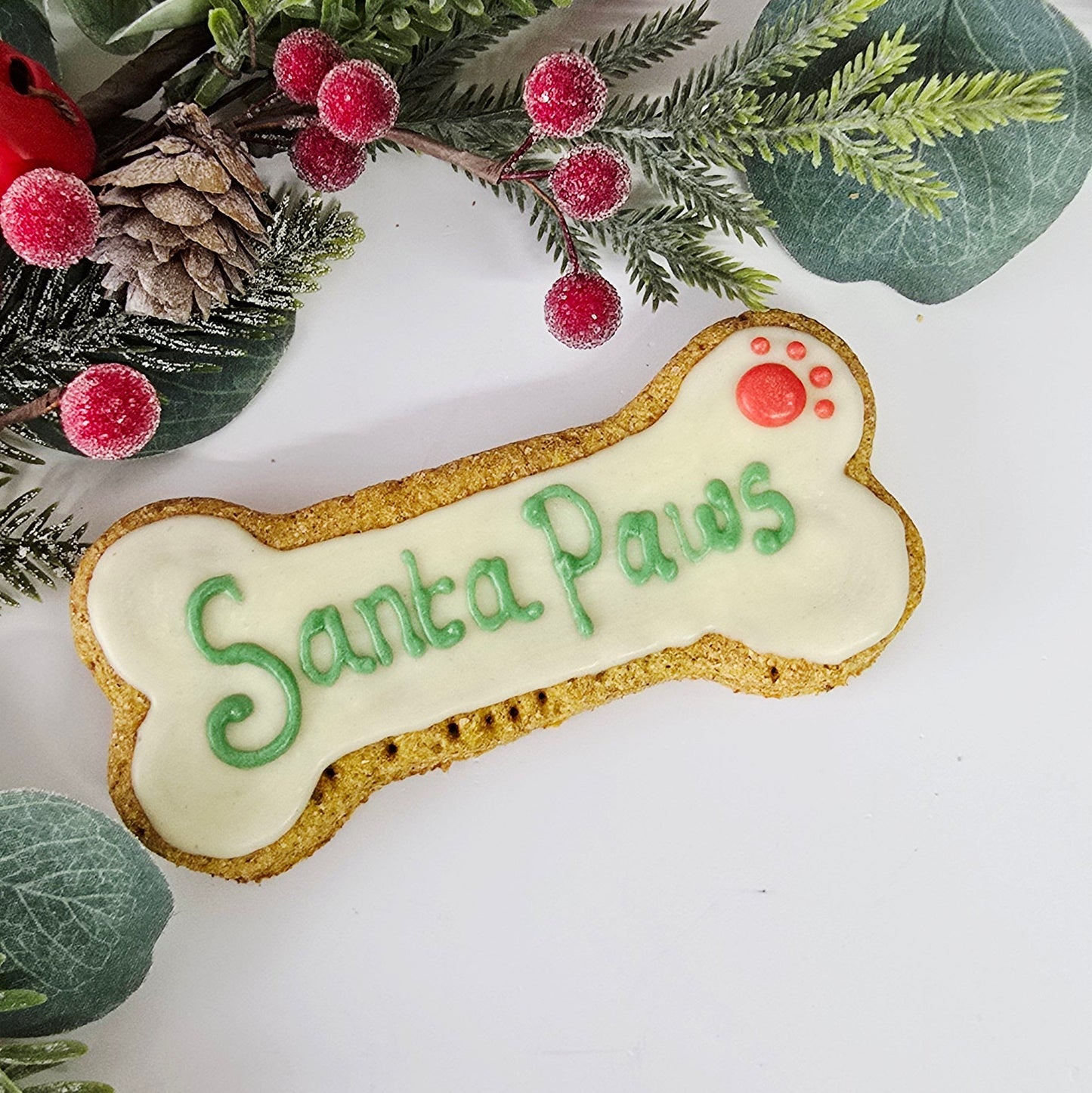 Dog bone-shaped cookie with 'Santa Paws' text surrounded by greenery on a white background