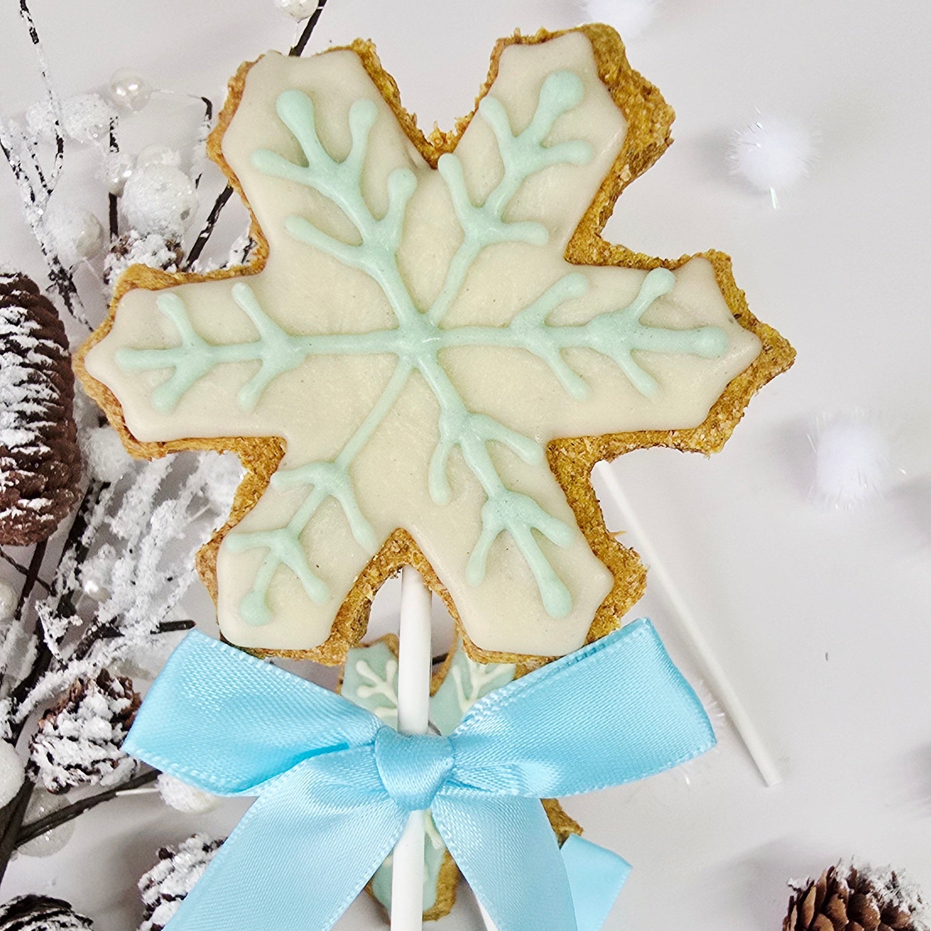 Snowflake-shaped cookie on a stick with a blue ribbon, surrounded by pine cones and branches on a light background.