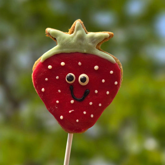Cookie shaped like a strawberry with a face on a stick against a blurred green background