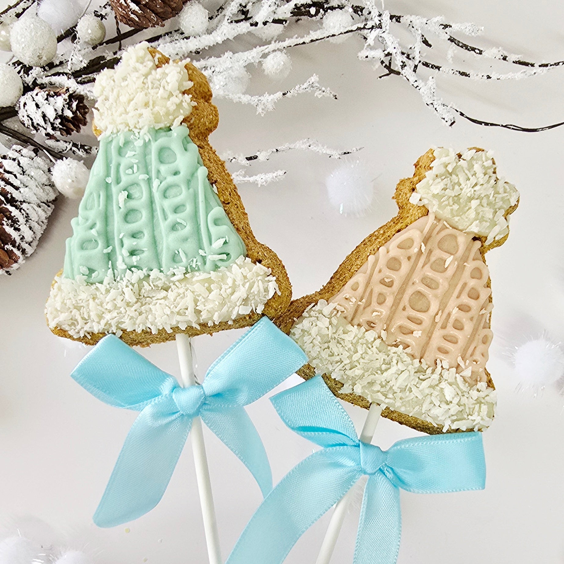 Two cookies on sticks shaped like people wearing knitted sweaters with blue ribbons against a snowy background.